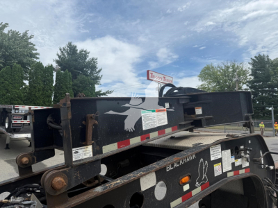Beam trailer frame with hydraulic beams and warning decals. Visible labels include 'BLACKHAWK' and safety stickers on a lowboy trailer.