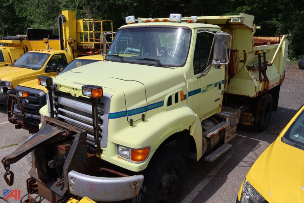 Dump truck in a fleet lot showing a Sterling L8500-style cab with a plow attachment and dump body. Visible text includes no obvious model numbers on the image; truck is pale green with blue stripe.