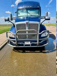 Kenworth T680 front view showing large grille, bull bar, and Texas license plate. Blue cab with windshield and side mirrors, water on ground.