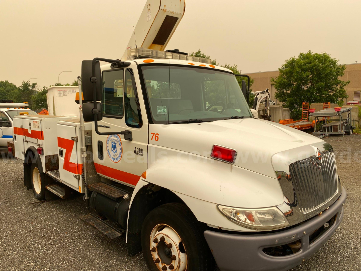 International 4300 bucket truck with a boom lift, white with orange stripe, showing side text and numbers on cab and body.