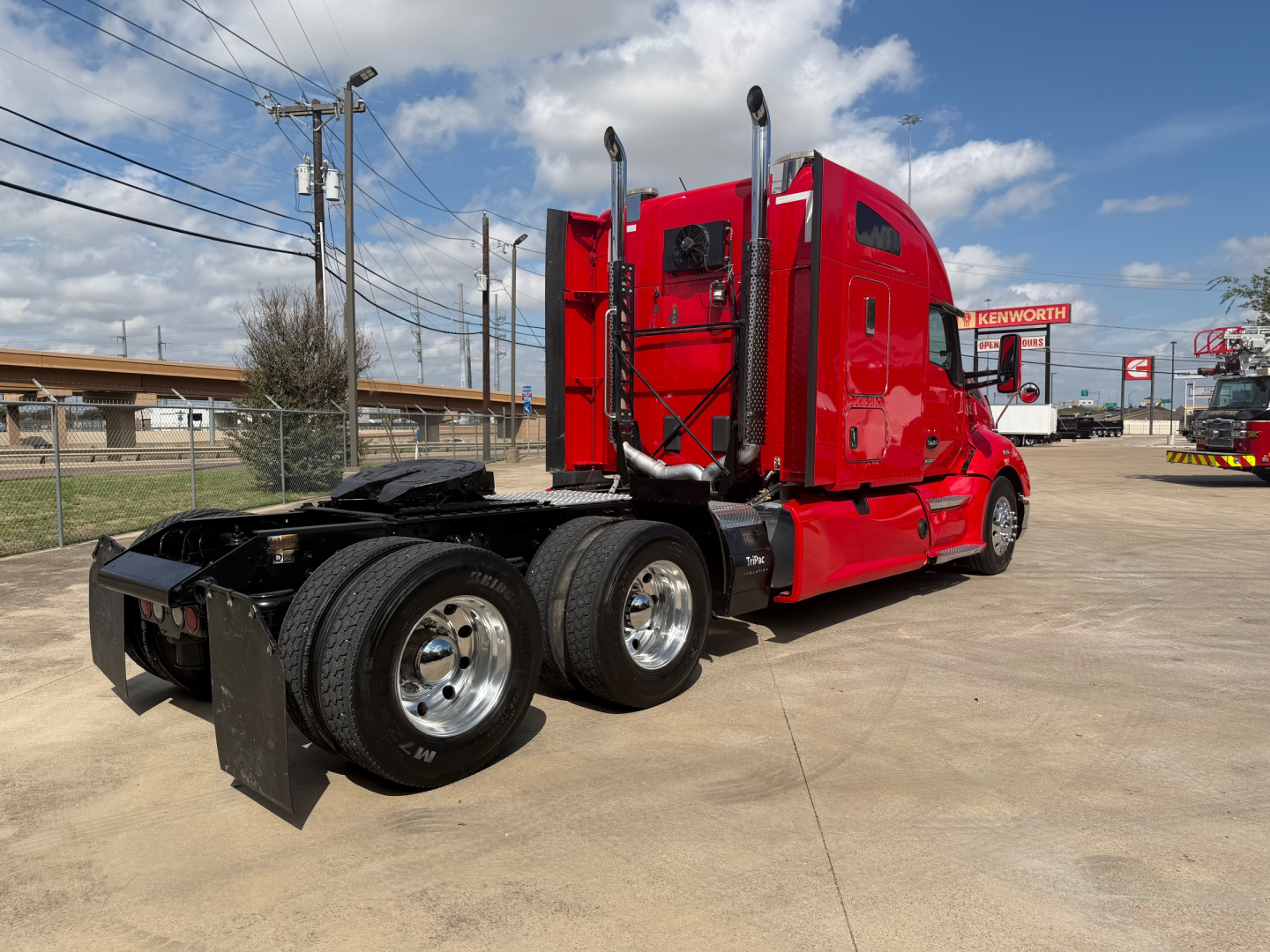 Kenworth T680 tractor with red cab, three rear axles, chrome wheels, exhaust stacks, and black mud flaps. Visible text includes Kenworth branding on sleeper and truck in distance.