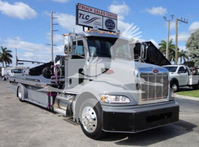 Peterbilt 337 rollback tow truck in front of dealership, chrome grille, side crane, and flatbed with wheels visible.