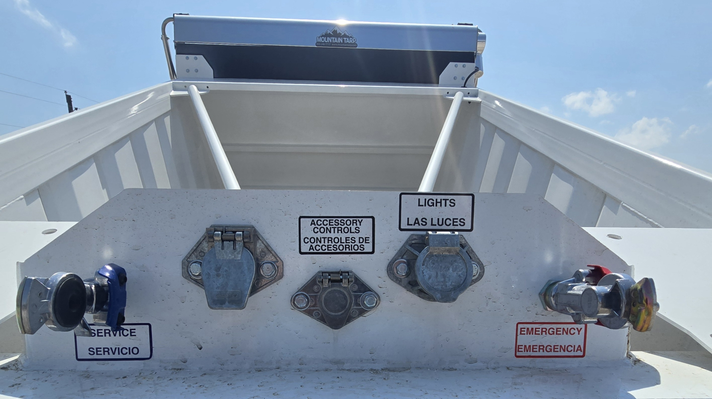Dump trailer rear with control levers and warning labels: service, emergency, lights, accessories controls; blue and red ports visible; metallic surface.