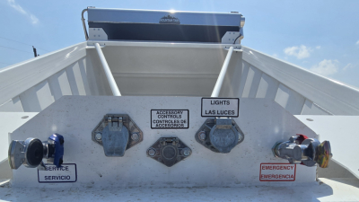 Dump trailer rear with control levers and warning labels: service, emergency, lights, accessories controls; blue and red ports visible; metallic surface.