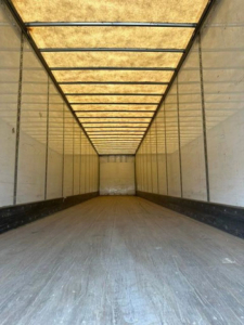 Interior of a van trailer viewed from the rear with long rectangular cargo space, light panels on ceiling, side walls, and empty plywood floor.