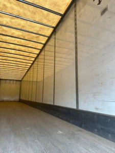 Trailer interior showing long empty cargo area with wooden floor and sidewalls, light-colored translucent roof and vertical support tracks.
