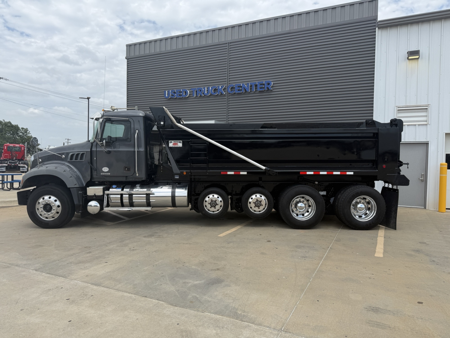 Dump truck showing a Mack GR64F dump truck with black body and multiple rear axles parked in front of a building labeled USED TRUCK CENTER.