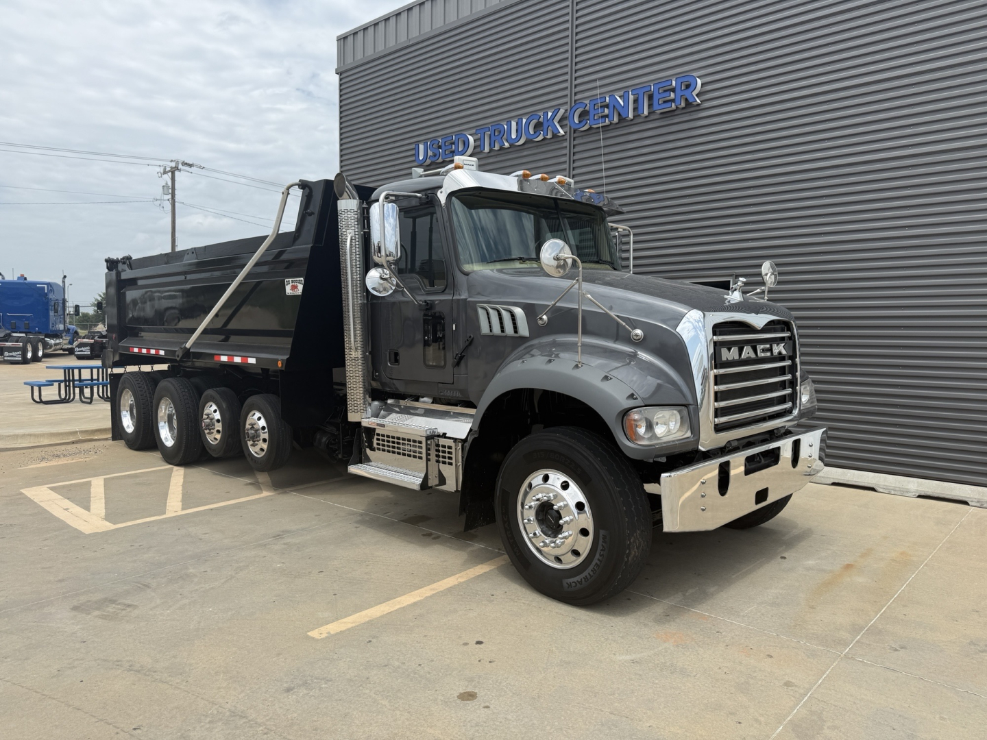 Mack dump truck in blue/gray showing Mack grille and chrome bumper. The truck has a black dump bed with six rear wheels. Visible text on the truck includes 'MACK' on the grille.