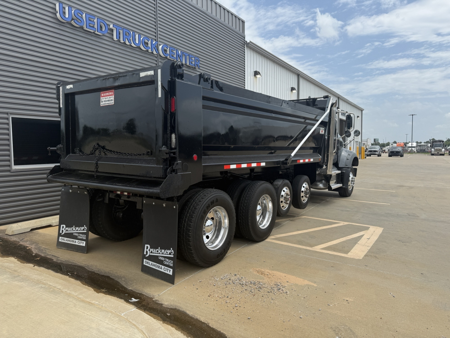 Dump truck side view showing black Mack GR64F dump body and triple rear axles with mud flaps near a dealer building.