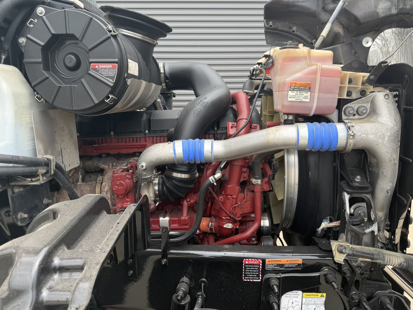 Mack GR64F engine bay showing red diesel engine, turbocharger, intercooler piping with blue silicone hoses, cooling reservoir, air intake assembly, and various hoses.