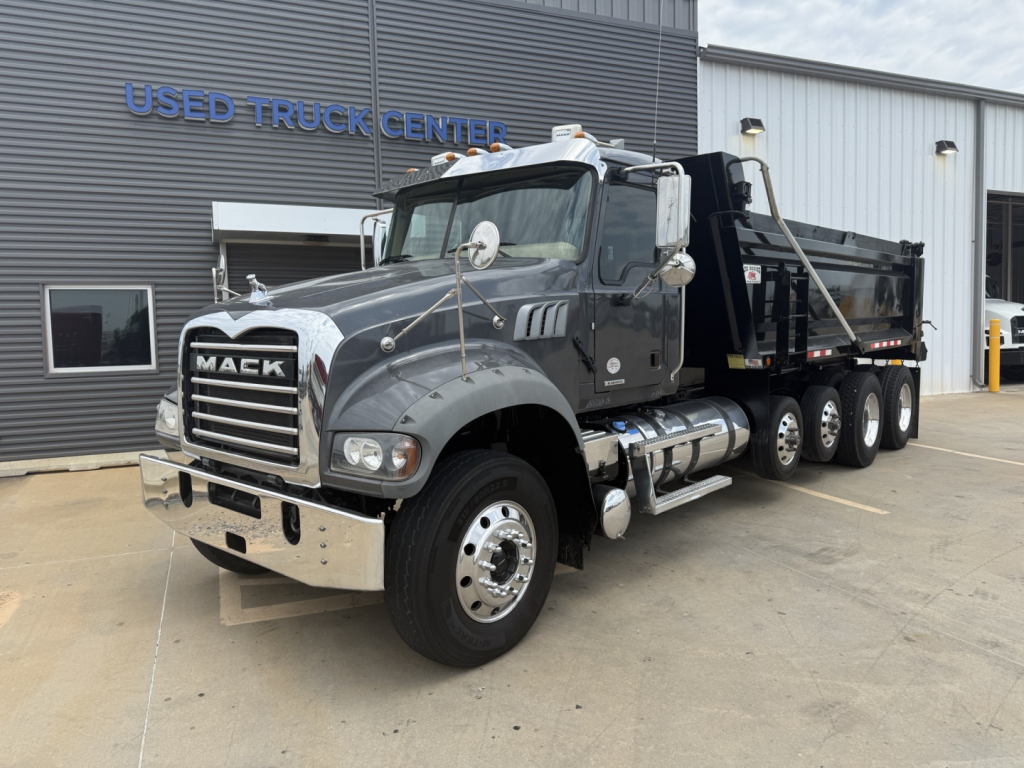 Truck: 2023 Mack GR64F dump truck with grey cab and black dump bed, front chrome bumper, visible Mack grille, triple rear axles, parked outside used truck center.