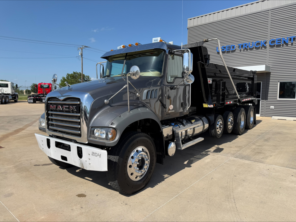 Mack GR64F dump truck in a dealership lot showing a gray cab, chrome mirrors, six rear axles, and a black dump bed.