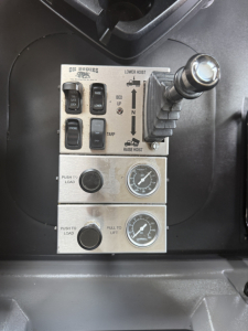 Control panels and joystick in Mack GR64F cab showing levers labeled bed, tare, raise hoist; switches with labels like lower hoist, push to load, pull to lift; gauges with needles.