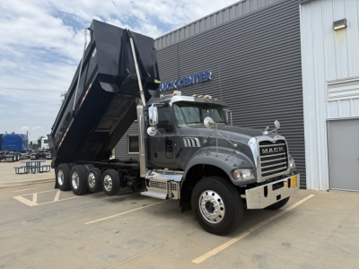 Mack GR64F dump truck with raised dump bed in front of a depot, chrome grille, dual stacks, and heavy-duty wheels.
