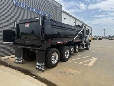 Dump truck side view showing black Mack GR64F dump body and triple rear axles with mud flaps near a dealer building.