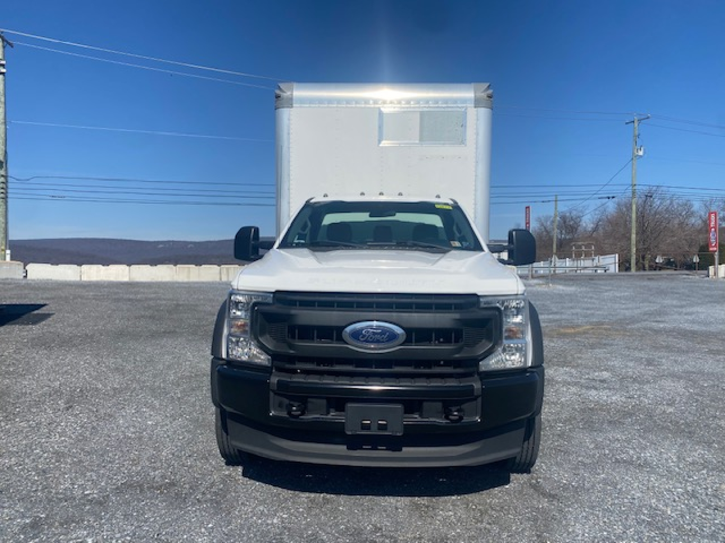 Ford F-550 box truck front view showing grille with Ford emblem, large side mirrors, and cargo box rear. Clear sky, vehicle on gravel lot.