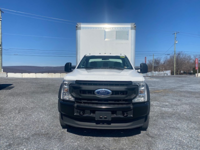 Ford F-550 box truck front view showing grille with Ford emblem, large side mirrors, and cargo box rear. Clear sky, vehicle on gravel lot.