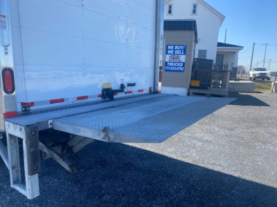 Ford F-550 box truck tail with loading ramp extended and rear chassis, silver metal platform, sliding ramp hinge near bottom left, gravel ground.