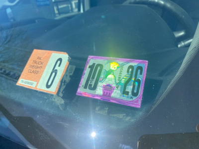 Vehicle windshield showing three inspection/tax stickers: orange 6 weight class label and purple 26 plate with green 10 overlay, on dash of a Ford F550 box truck.