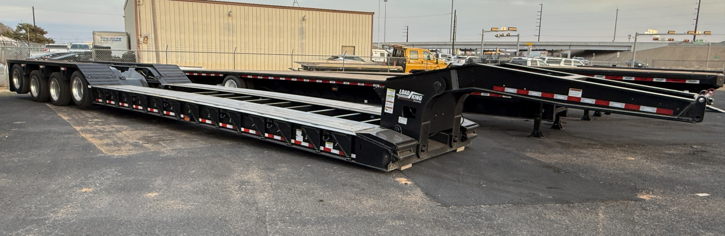 Load King 554-Quad 9' Wide 24'LDH trailer in yard, lowboy configuration with multi-axle extendable deck and landing gear, long black frames with reflective safety tape.