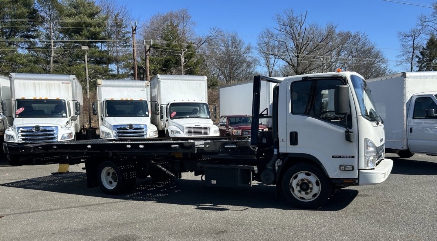 Isuzu NRR tow truck/flatbed vehicle side view showing box vans in background