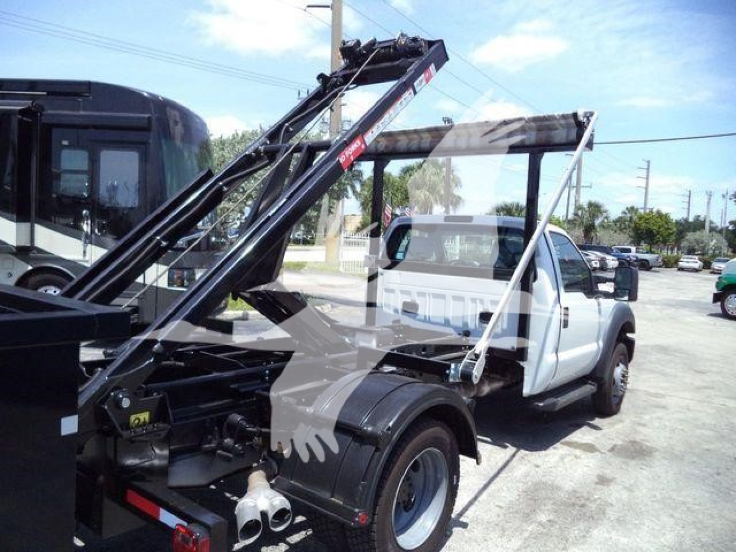 Truck with a roll-off hoist in folded position, white cab, black hydraulic boom arms, viewed from the driver's side rear.