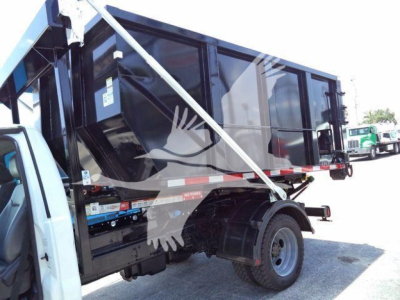 Truck bed of a 2016 Ford F550 roll-off body showing side hydraulic lines and rear wheel, with reflection of other trucks in the background.