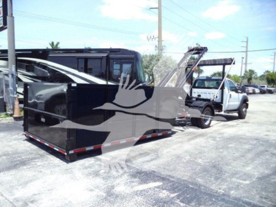 Flatbed tow truck with black enclosed bed and a white pickup tow vehicle in rear, dealership lot in background.