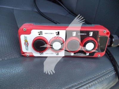 Control pendant with red housing and three round push buttons, labels show arrows and icons; text includes 'SC...', 'S W...' and icons; device lies on gray seat with black cable.