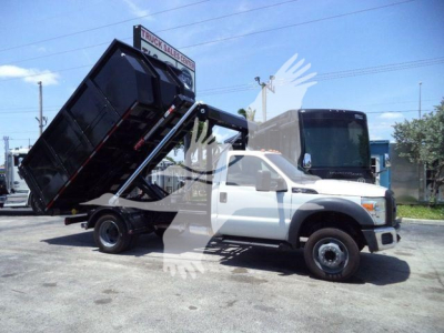 Ford F-550 regular cab chassis with a raised black roll-off bed in a dumping position.