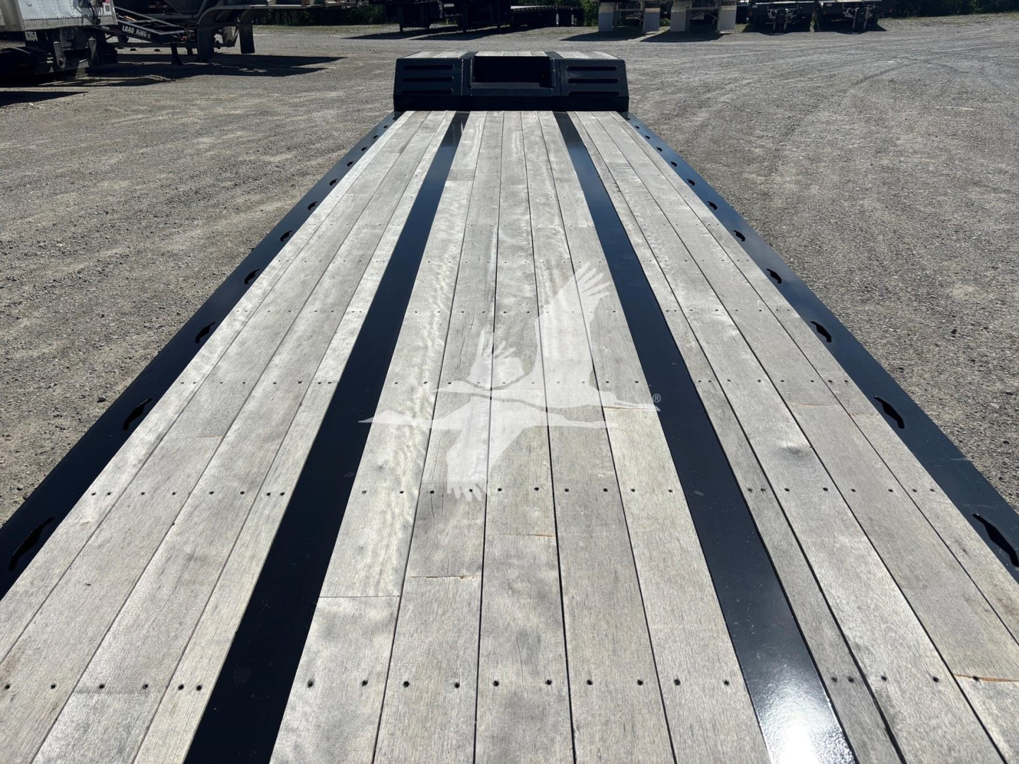 Flatbed trailer deck with wooden planks and black steel rub rails, view from front with gooseneck area in distance