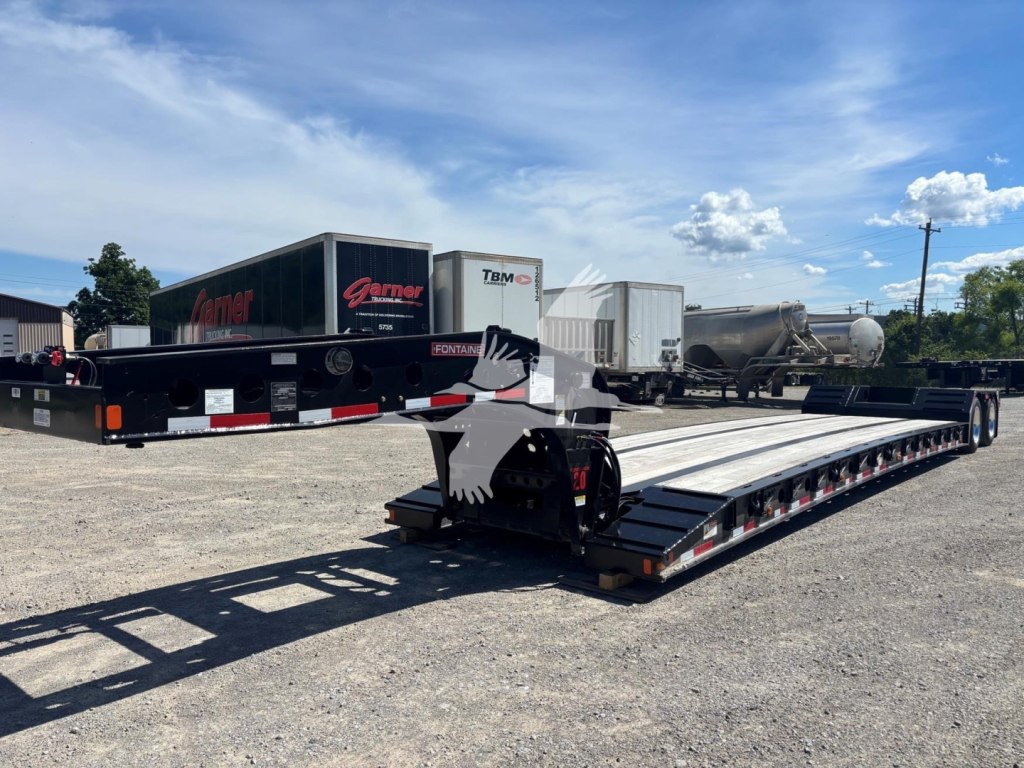 Fontaine Renegade lowboy trailer in a parking lot showing long flatbed with low deck and gooseneck; rear section visible with tandem axles and loading ramps.