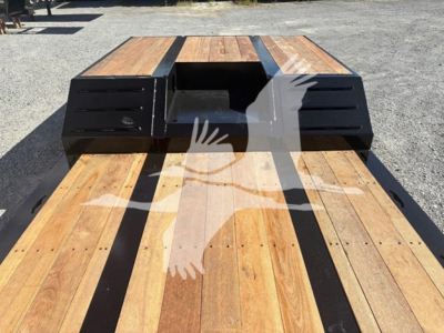Flatbed trailer deck with wooden planks and black toolbox housings on each side, open center cavity near cab end.