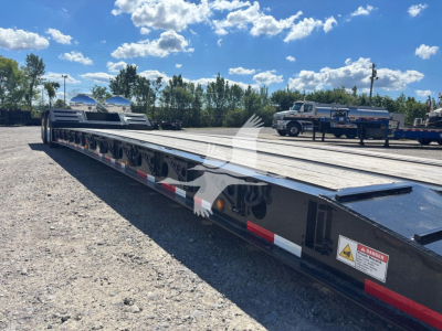 Lowboy trailer on a yard with clear sky; Fontaine Renegade N20 flatbed with long deck, multiple axles, reflective tape along the sides, warning label near front.