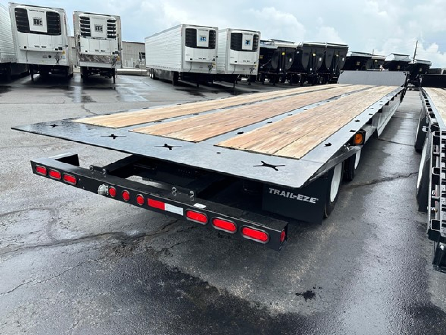 Trail-Eze TE801 trailer with wooden deck on a flatbed, rear view showing taillights and ramps, on a wet lot with other trailers in background.