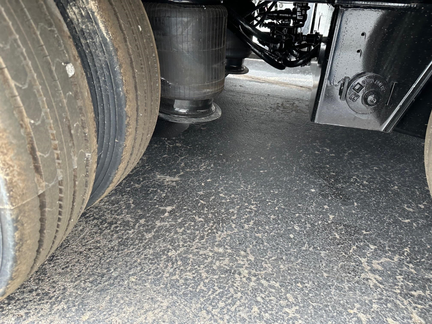 Trailer undercarriage showing dual tires, hitch area, and suspension components with gravel/dust on ground.