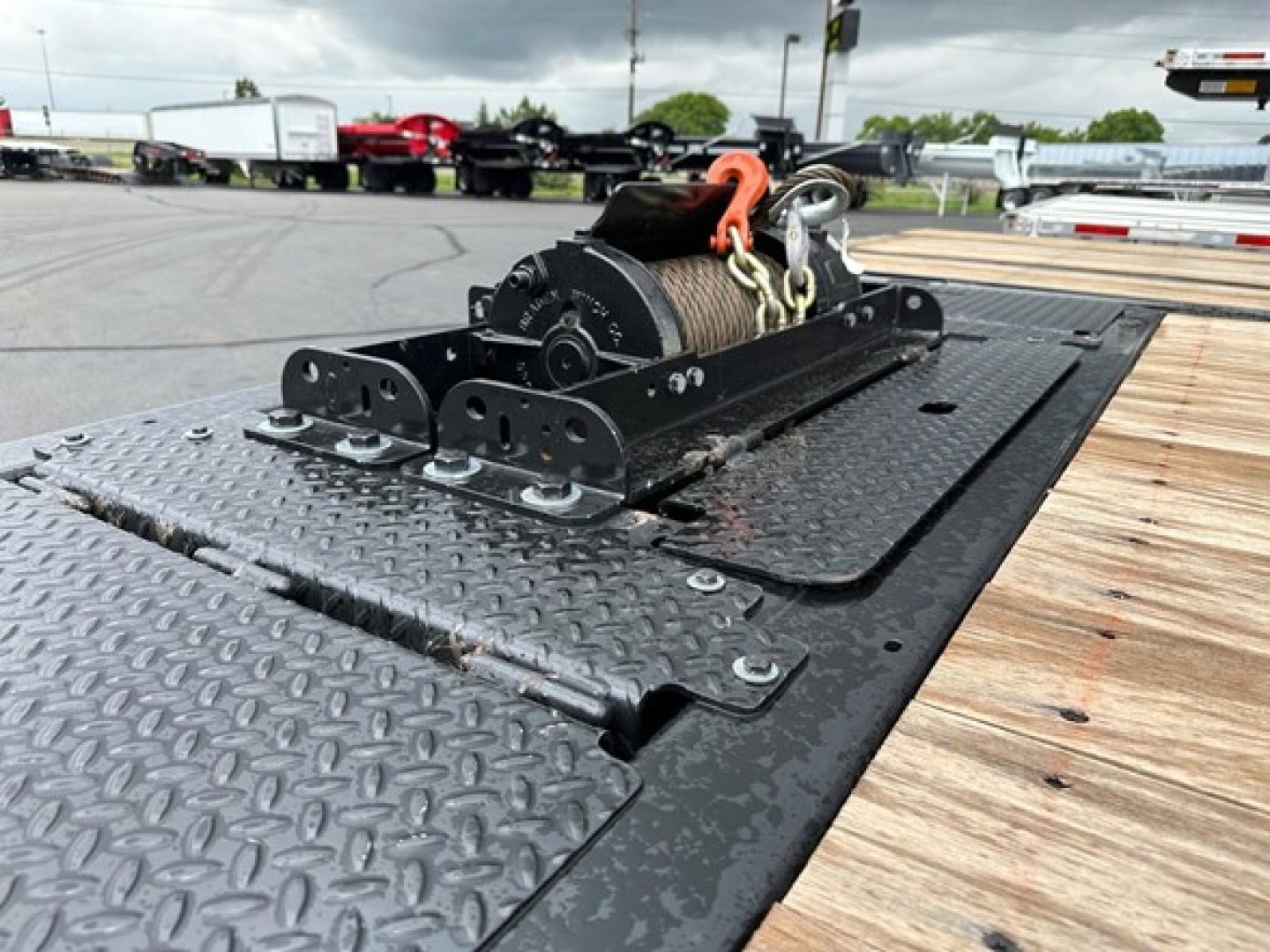 Winch-mounted hoist on a black diamond-plate platform with orange hook and steel cable; trailer deck and part of road in background.