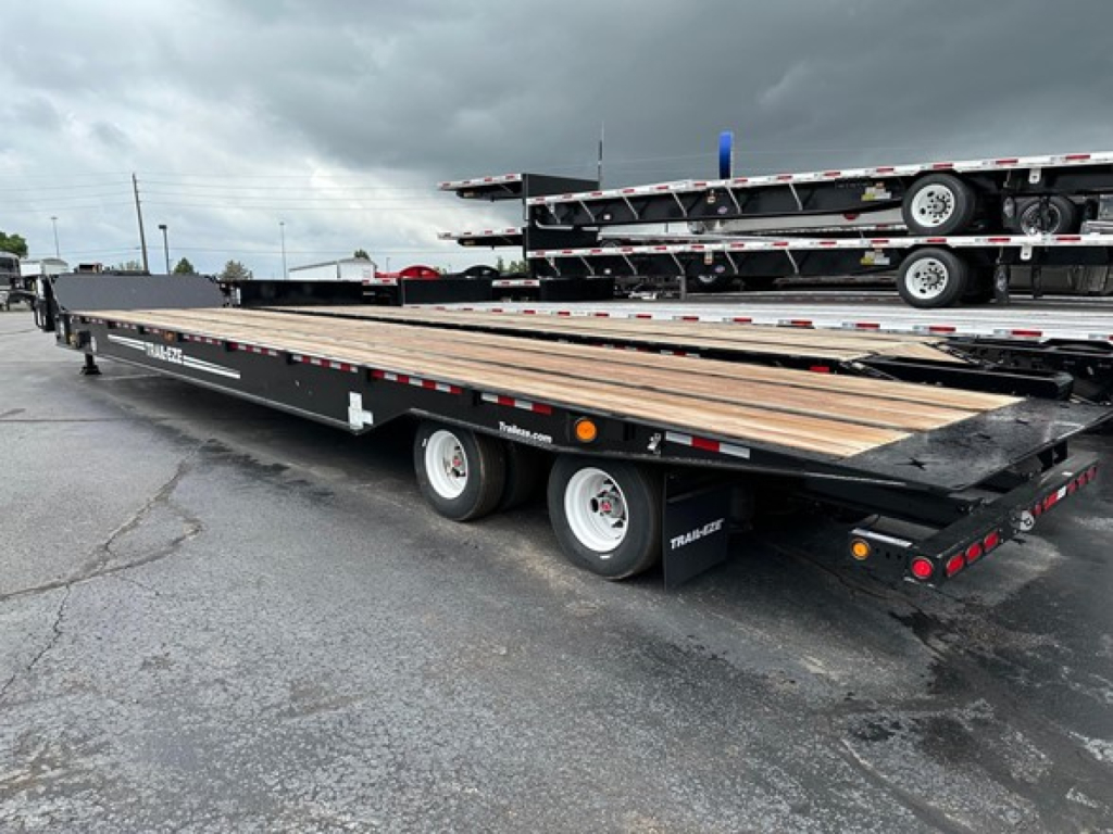 Trailer shows flatbed trailer with long wood deck and dual axles, left side branding reads Trail-Eze, NE9777 stock, flatbed equipment visible in background.