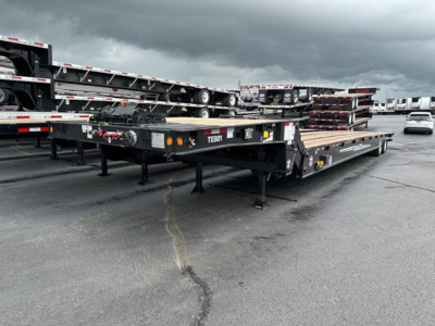 Flatbed trailer in a dealership lot showing TE801 model plate and rear deck with ramps, highway overcast sky.