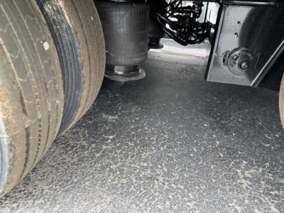 Underneath a trailer showing part of a wheel, suspension components, and chassis with dirt on the ground.