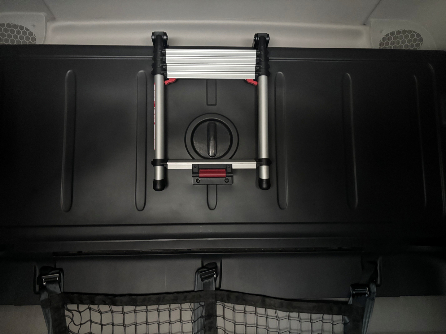 Truck cabin interior showing a compact folding ladder mounted on a black surface, with a circular cap and red latch beneath. Spare wheel storage area visible.