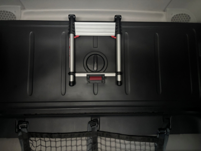 Truck cabin interior showing a compact folding ladder mounted on a black surface, with a circular cap and red latch beneath. Spare wheel storage area visible.