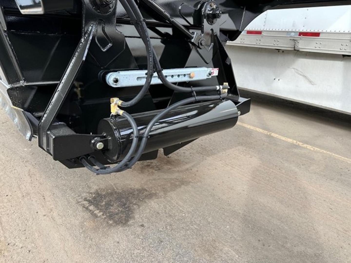 Trailer hitch area showing hydraulic mechanism and support cylinder on a bottom-dump trailer. Visible components include hydraulic lines, a black cylindrical hydraulic cylinder, mounting brackets, and a silver support arm.