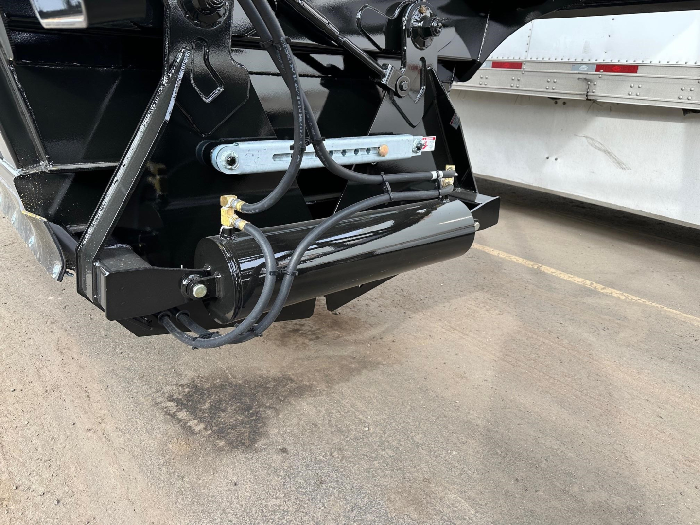 Bottom-dump trailer hitch area showing hydraulic cylinder and control linkage with hoses and black painted metal frame.