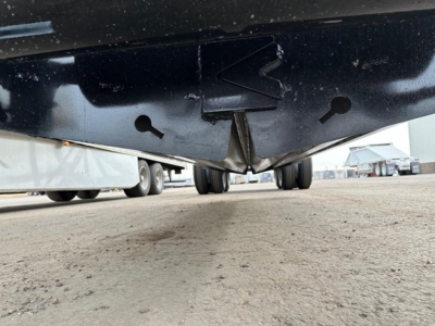 Under view of a trailer underside showing hitch/kingpin area with black metal and spare wheel hints in background.