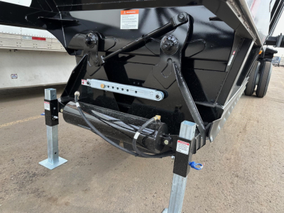 Bottom-dump trailer hitch area showing hydraulic cylinders and support legs with safety labels, structure in black and silver, wiring harness, and trailer frame.
