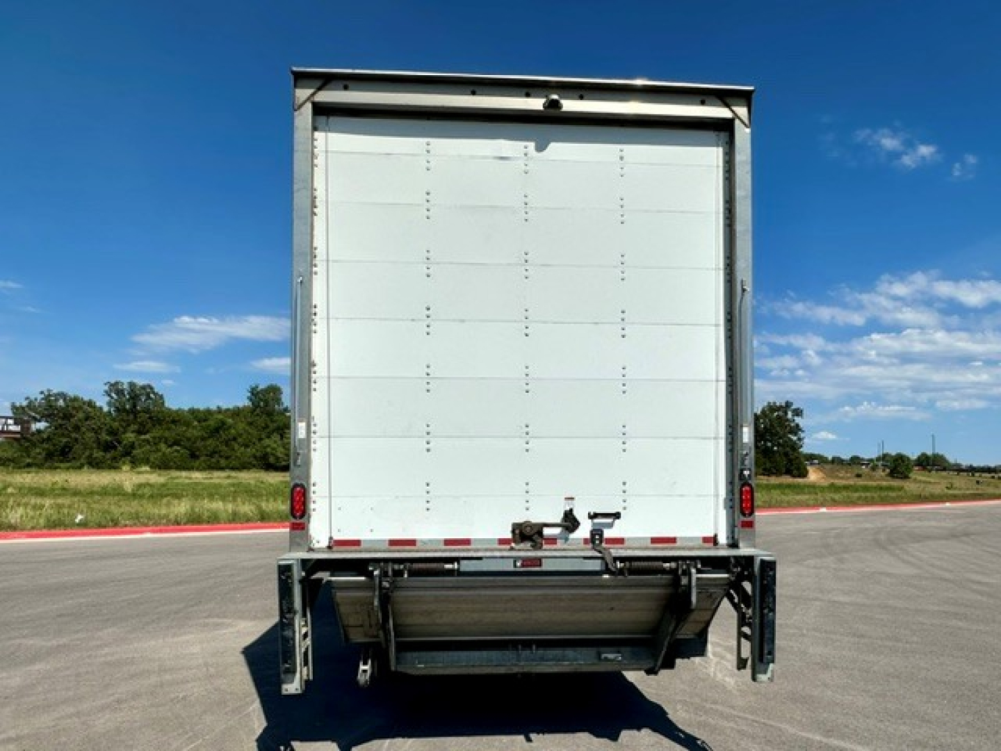 Freightliner M2-106 rear view showing roll-up box truck with raised tailgate and reflective rear bumper, clear sky, open road.
