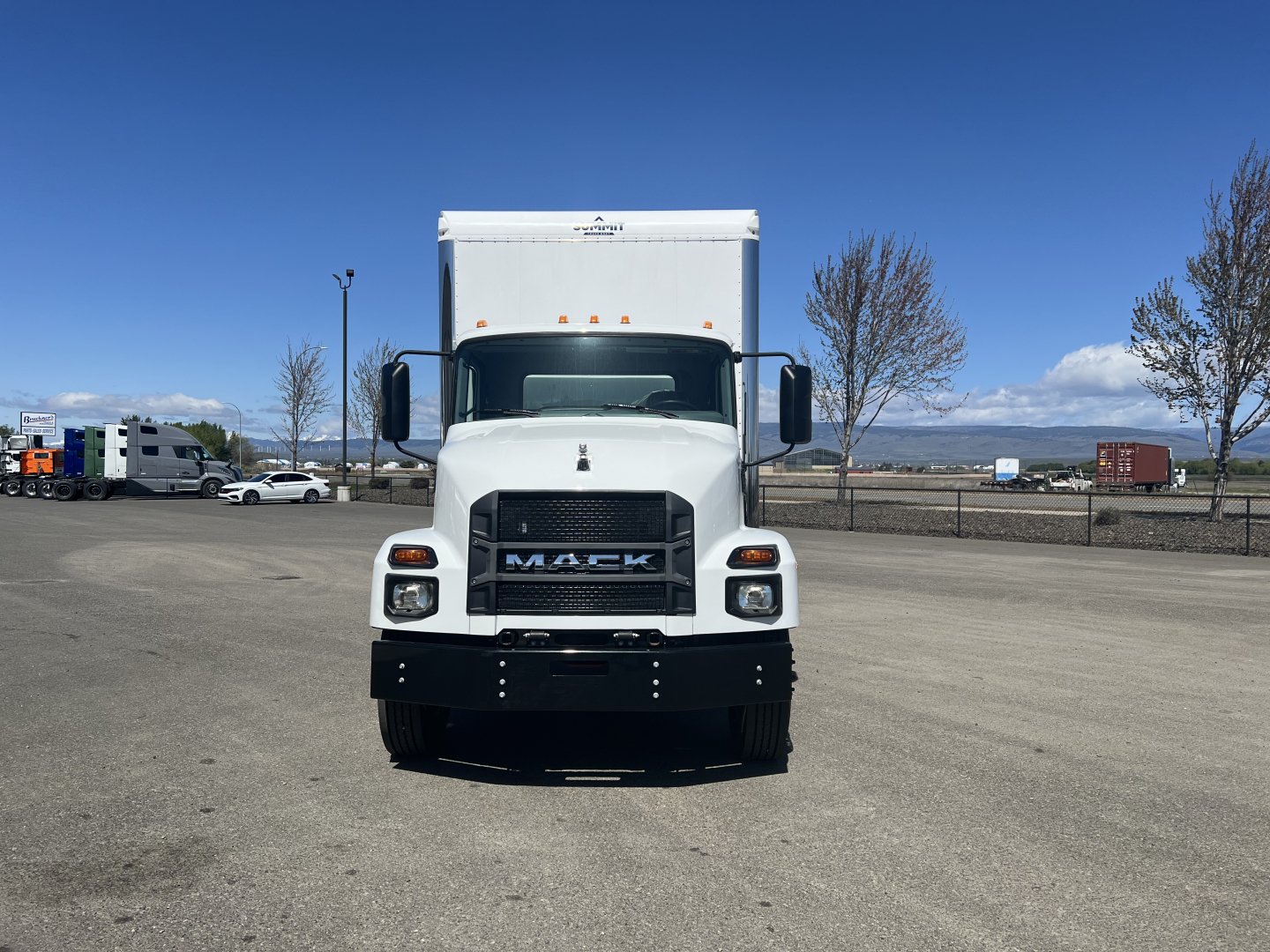Front view of a white 2025 Mack MD6 box truck showing grille, bumper, headlights, and cargo box; clear blue sky and parking lot in background.