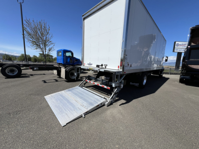 Box truck with rear aluminum ramp lowered, white cargo box, blue tractor in distance. Visible text: no legible labels on box exterior. VIN plate not shown.