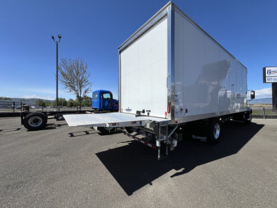 Box truck with a large white cargo box and a fold-down rear ramp, viewed from the passenger side rear, Mack MD6 trailer in a dealership lot.